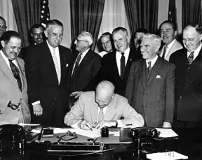 President Eisenhower seated at his desk signing a bill while a group of men in suits looks on, in black and white